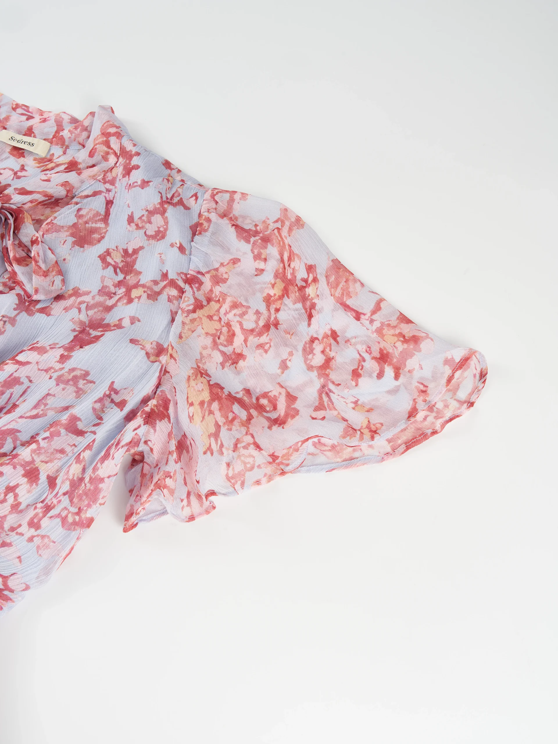 A lightweight floral mini dress featuring a feminine tie neckline, flutter sleeves, and a softly gathered waist. Its airy silhouette makes it ideal for summer collections, resort wear, and effortless everyday styling.