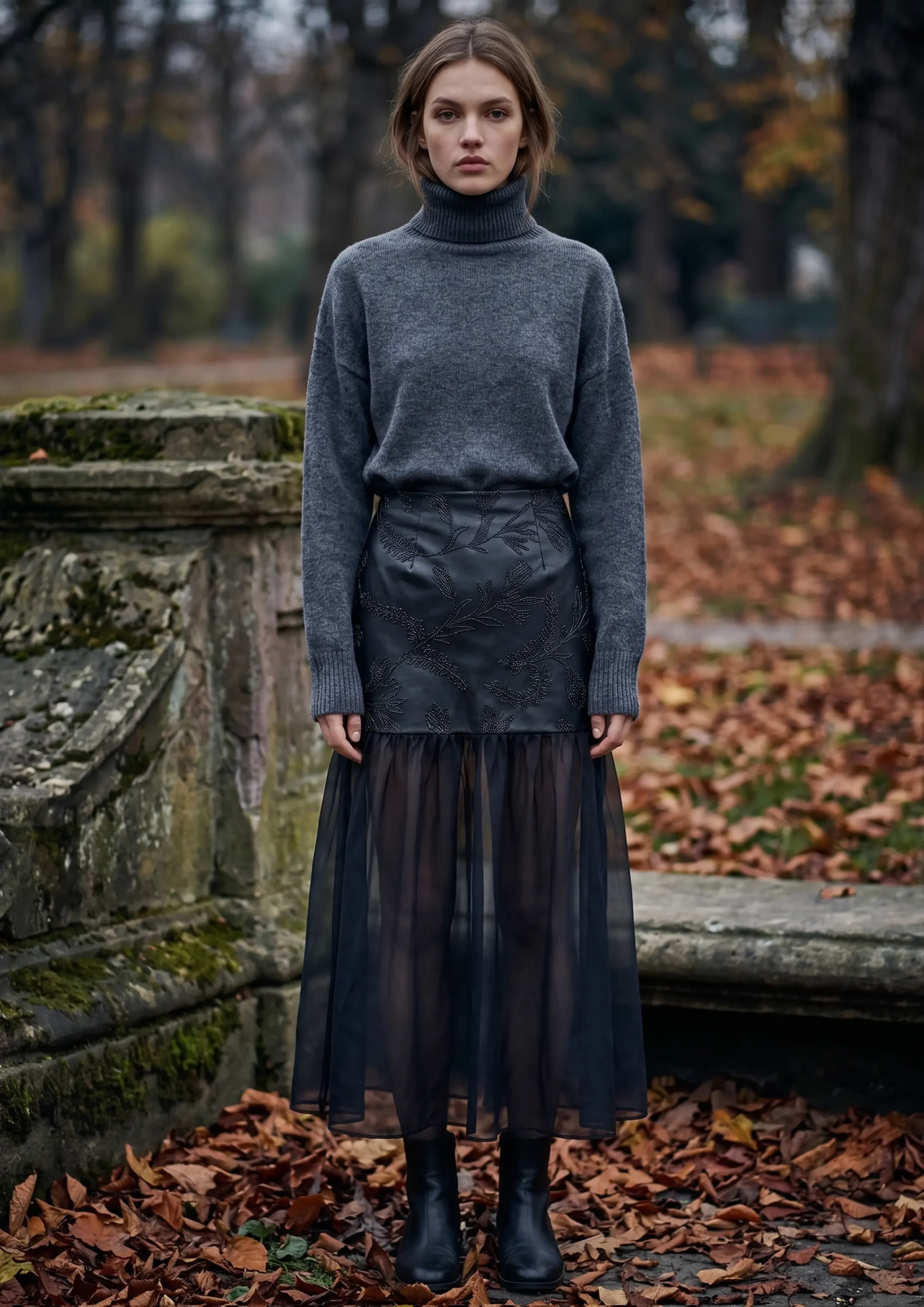 Woman standing outdoors in autumn wearing a gray turtleneck sweater and a black embroidered sheer-hem skirt with black boots, facing the camera with a neutral expression.